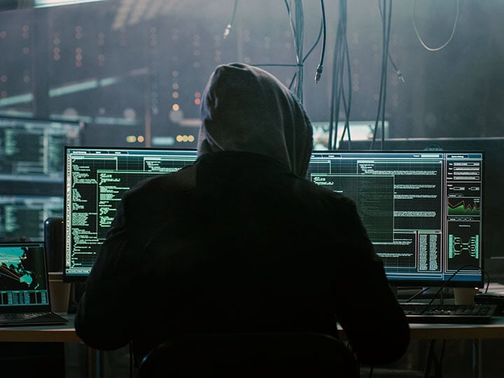 Hooded hacker in dark hideout on a computer with multiple display screens, breaks into data servers with a virus.