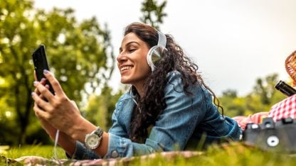 Woman lying on grass outdoors, smiling at her phone while wearing headphones.