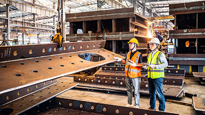Two male industrial workers standing in a steel factory looking at a large piece of steel that is hanging on a crane hook  